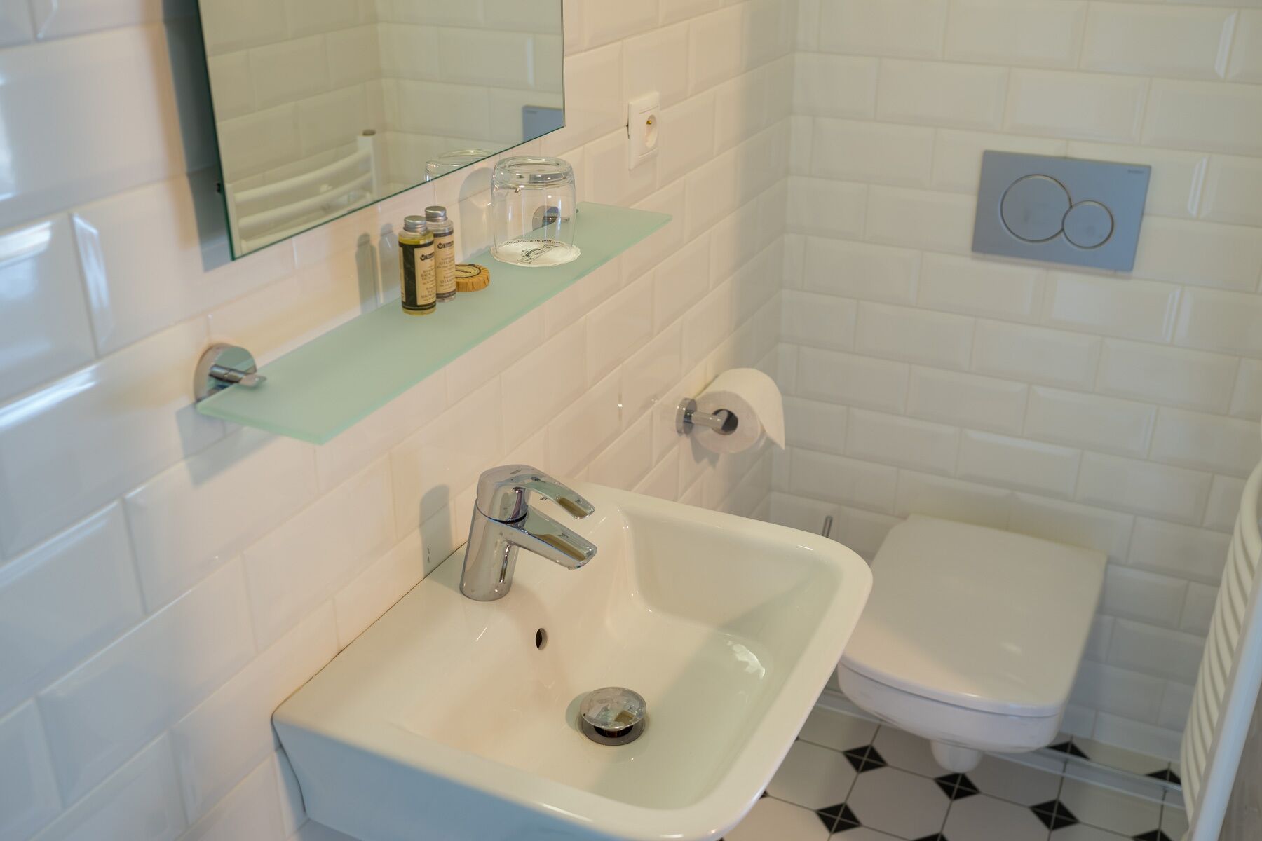 A bathroom with a white sink, a mirror, and a toilet. There is a glass shelf above the sink with toiletries.