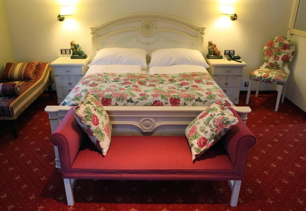 Una cama con ropa de cama floral, dos mesitas de noche con lámparas, un sillón tapizado con motivos florales y un suelo cubierto de alfombra roja.