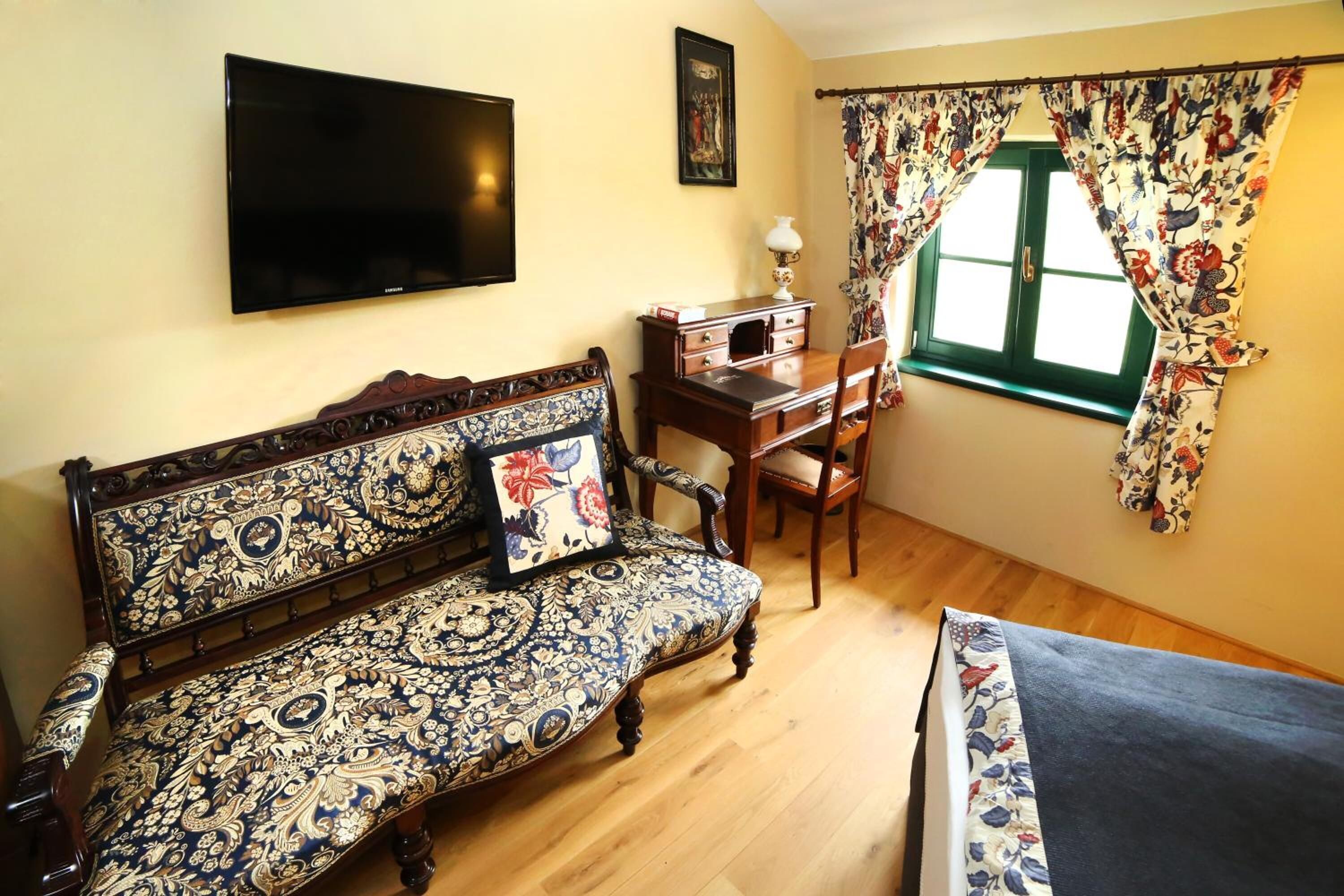 A room with a patterned sofa, a desk with a chair, a television mounted on the wall, and a window with floral curtains.