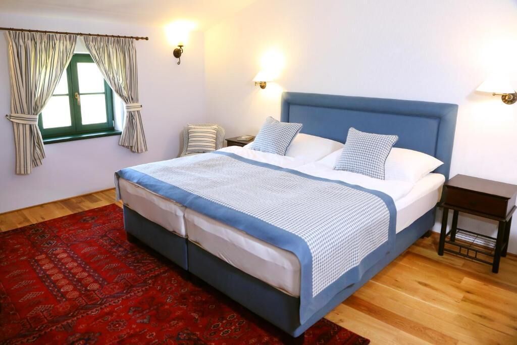 A bedroom with a bed, a nightstand, a lamp, a window with curtains, and a red carpet.