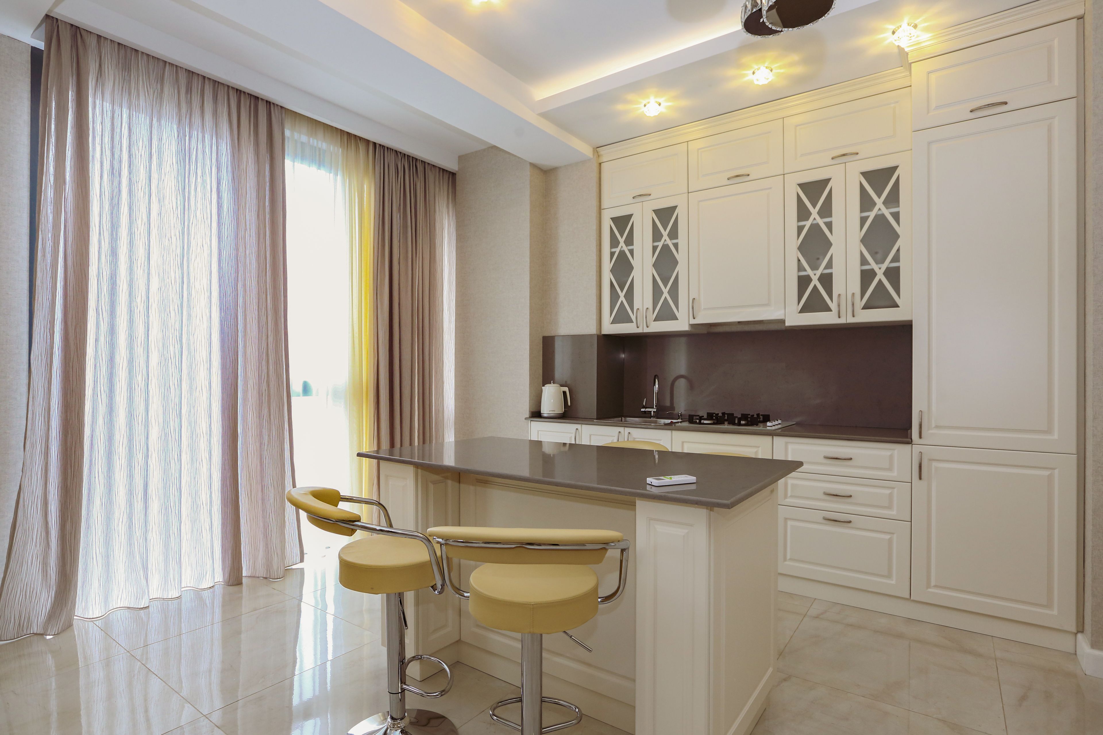 A modern kitchen with white cabinetry, a central island with bar stools, and a large window with curtains.