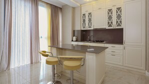 A modern kitchen with white cabinetry, a central island with bar stools, and a large window with curtains. - Hosthub - 2BR Super view Apartment (Tbilisi)