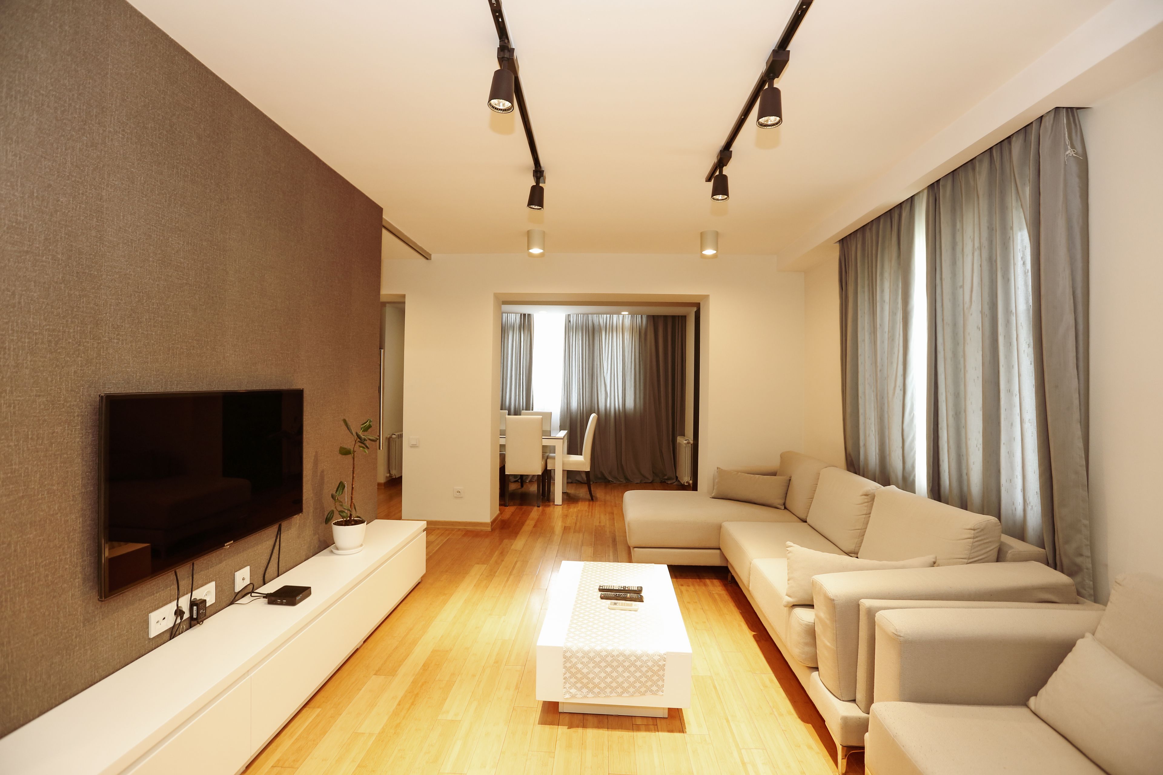 A modern living room with a flat-screen TV, a white console table, a beige sofa set, and a dining area in the background.