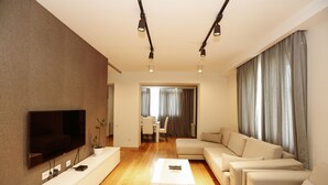 Una sala de estar moderna con un televisor de pantalla plana, una consola blanca, un juego de sofá color beige y un comedor al fondo.