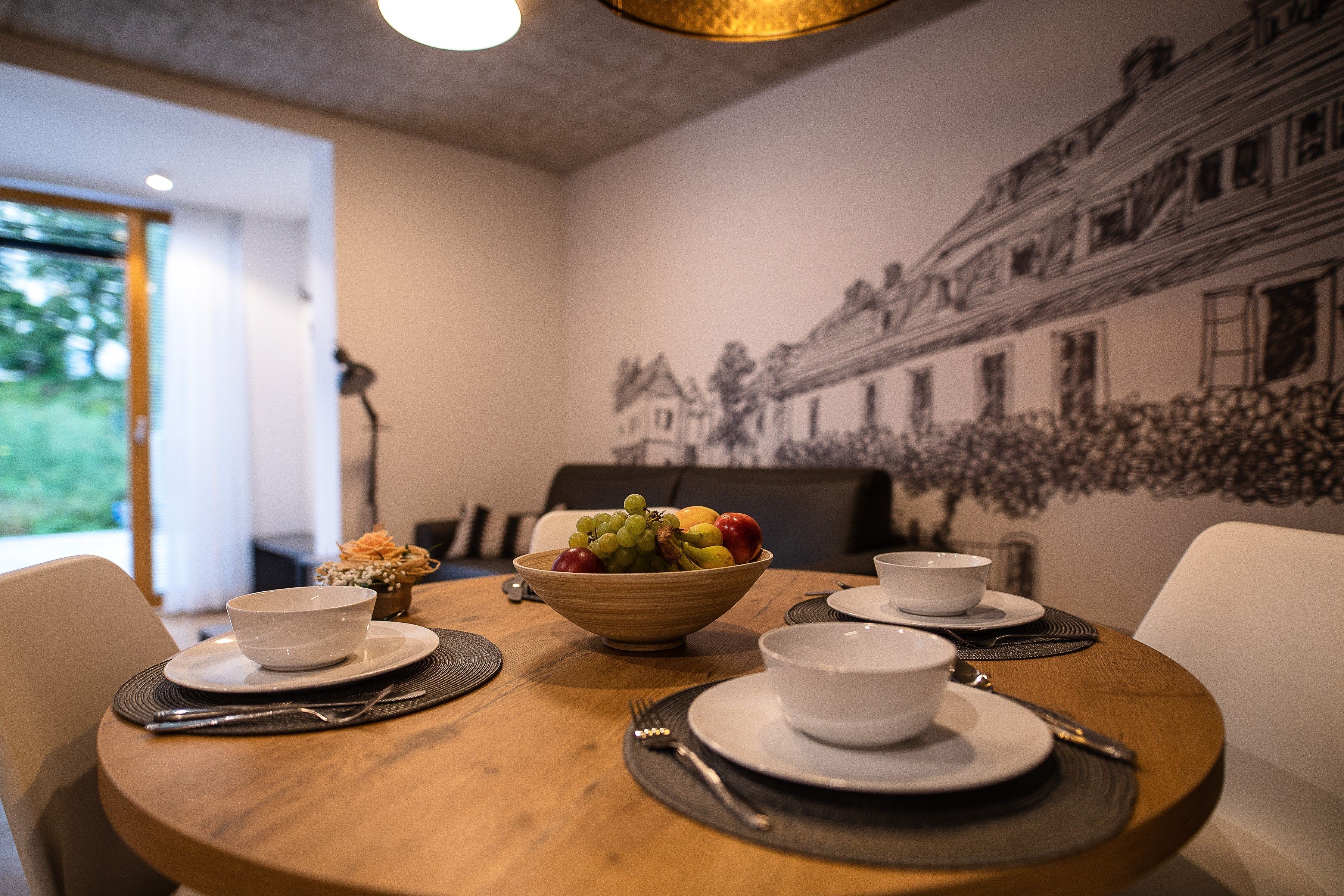A dining table set with white bowls, plates, and cutlery, a bowl of fruit, and a wall mural.