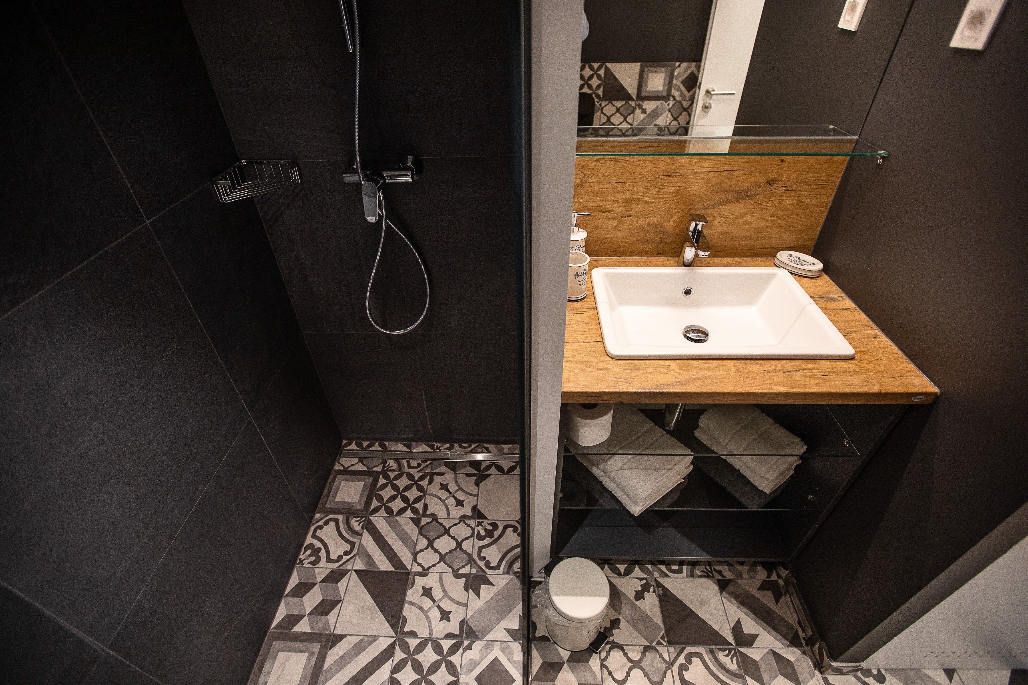 Un bany modern amb dutxa, un lavabo amb taulell de fusta, rajoles de terra amb disseny i una prestatgeria de vidre.