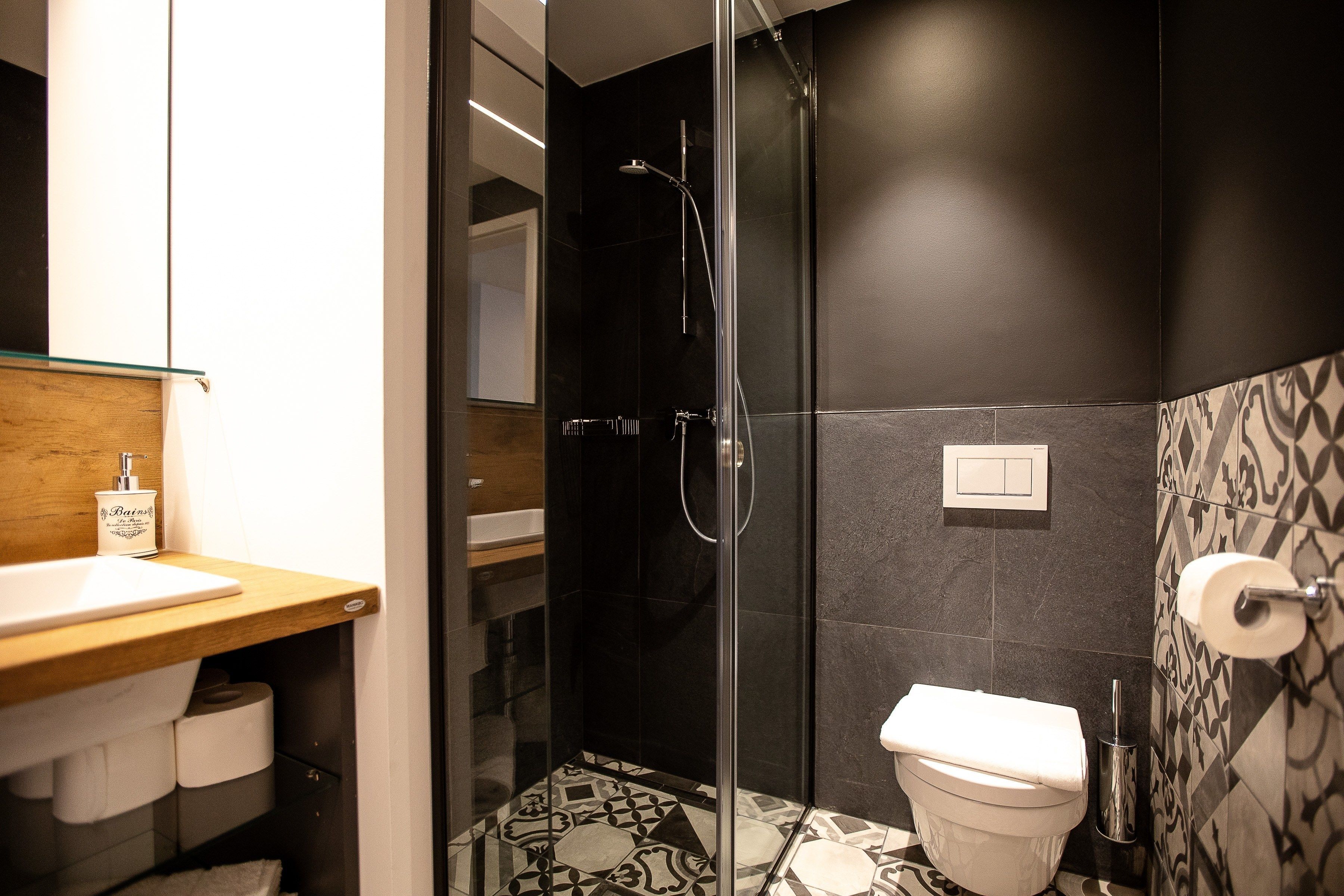 Un baño moderno con ducha envuelta en vidrio, tocador de madera con lavamanos y un inodoro con revestimiento de azulejos estampados.