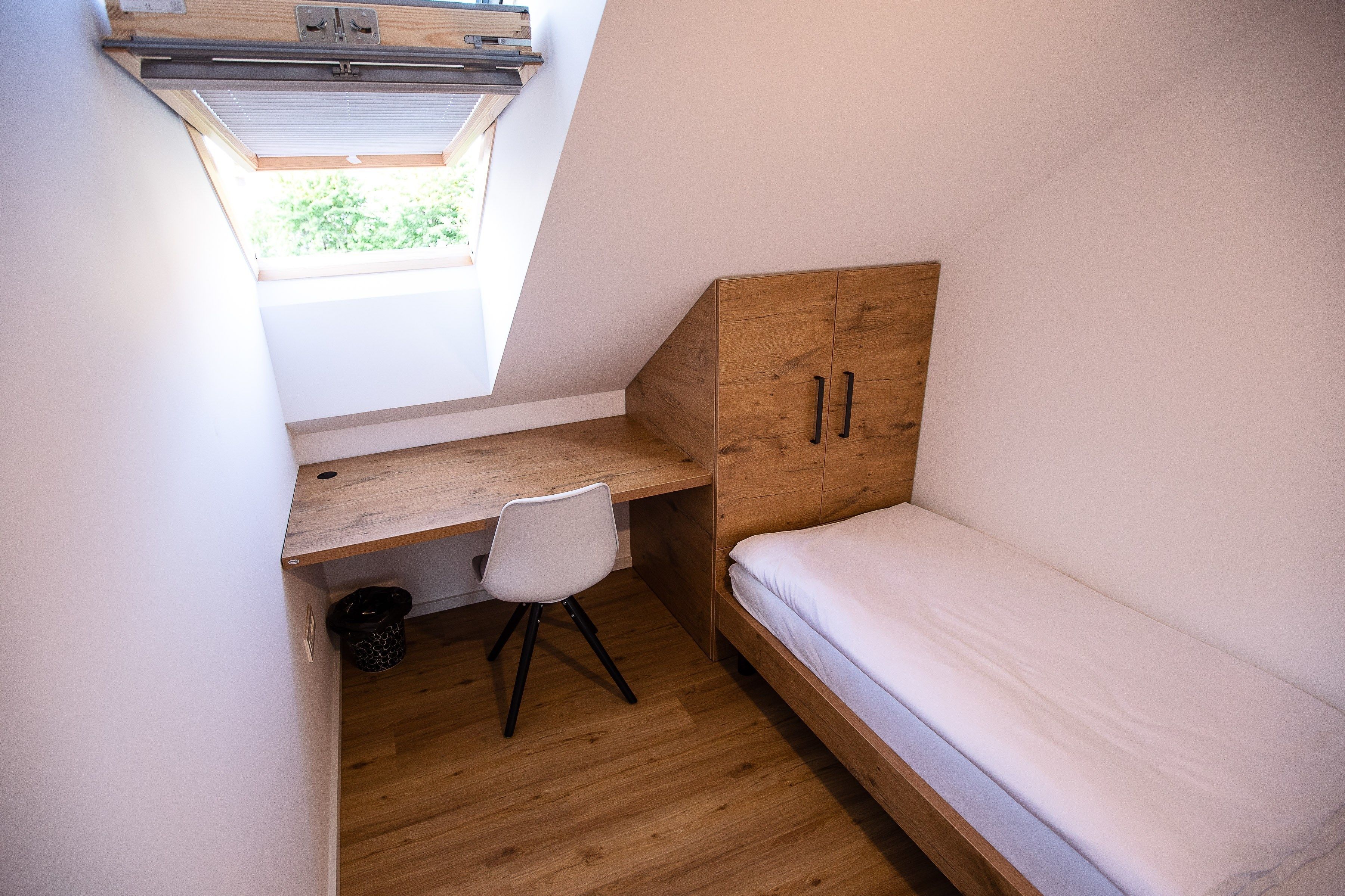 Una stanza compatta con travi a vista, una scrivania in legno, una sedia bianca e un letto singolo.