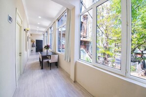 Un pasillo estrecho con una mesa de comedor y sillas, ventanas amplias con vistas a edificios y árboles, y una puerta al final.