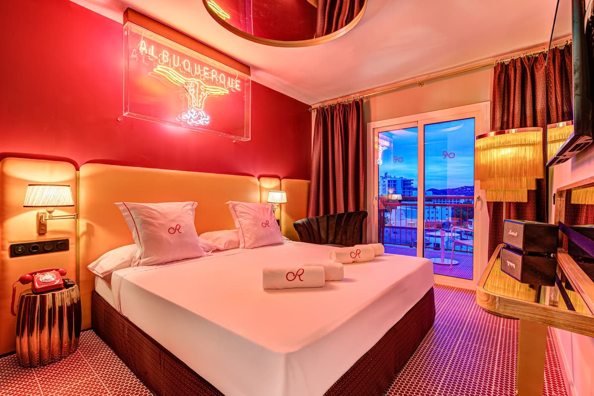 A hotel room with a bed, a telephone, a lamp, a table with a radio, and a view of the city through the window.