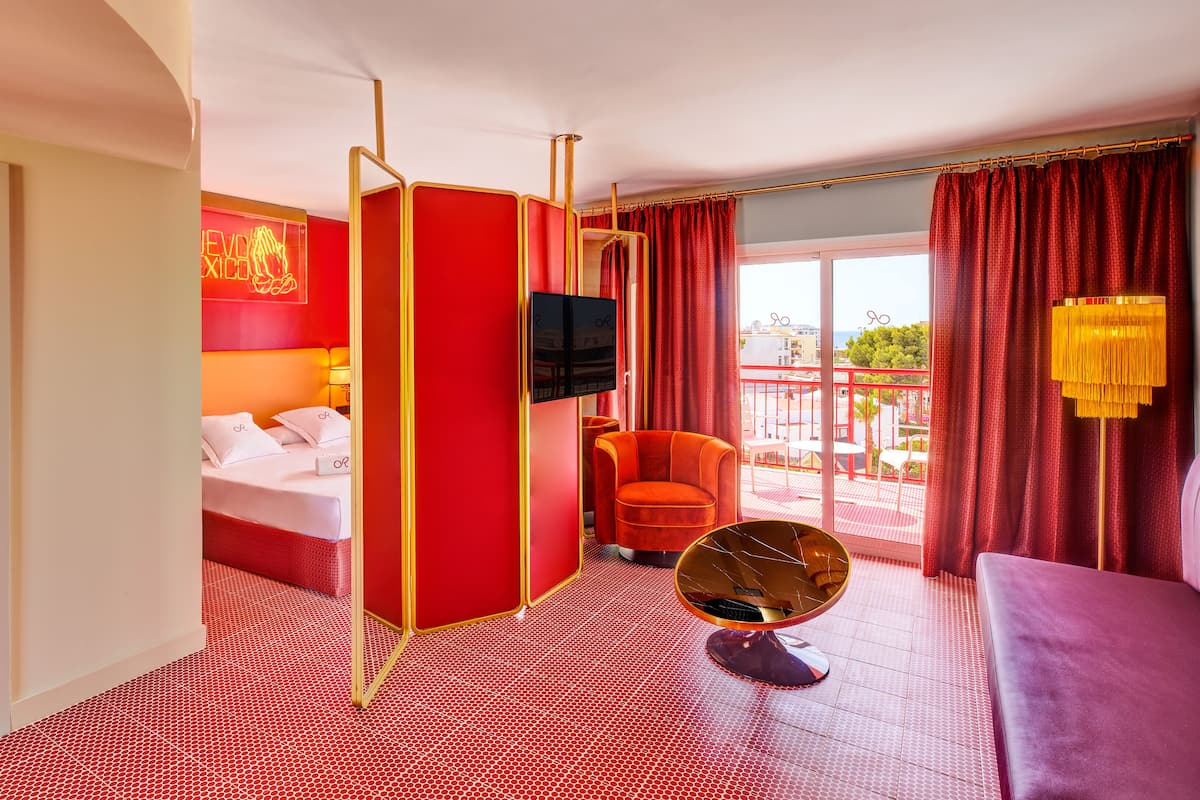 A hotel room with a red and white color scheme, a bed, a TV, a chair, and a balcony with red curtains.