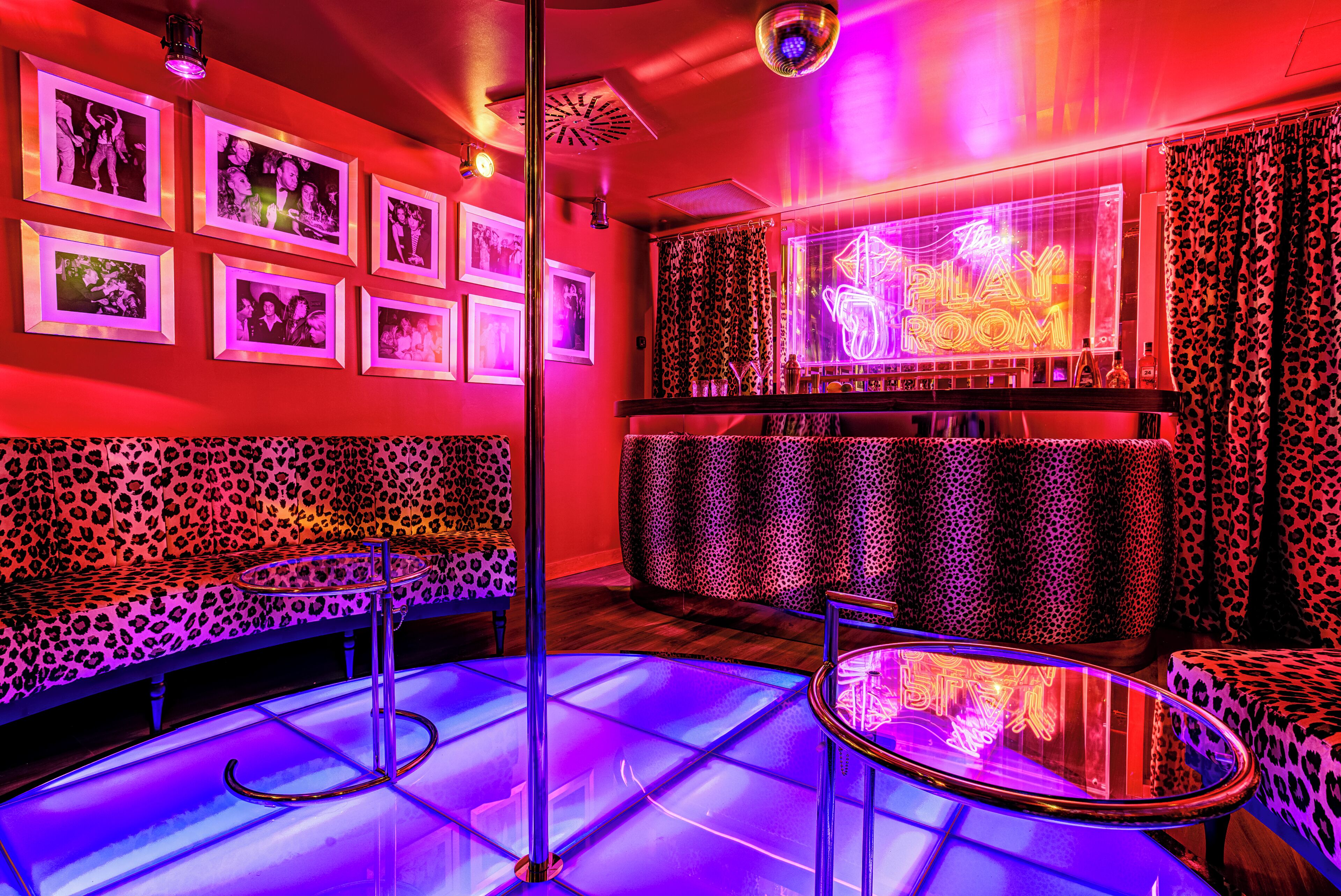 A room with a red and black leopard print sofa, a neon sign reading "The Play Room," and framed pictures on the wall.