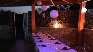 Uma área de refeições ao ar livre com uma mesa comprida posta para uma refeição, cercada por paredes de pedra e uma pérgola de madeira.