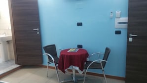 Quarto com parede azul, porta de madeira, pequena mesa com toalha vermelha, duas cadeiras e pia branca ao fundo.