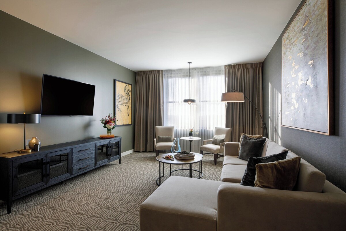 A modern hotel room with a flat-screen TV, a sofa, a coffee table, and a dining area with chairs.