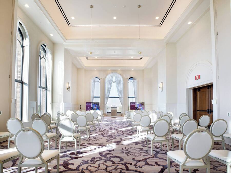 Un salón de conferencias con filas de sillas blancas, ventanas amplias y una alfombra estampada.