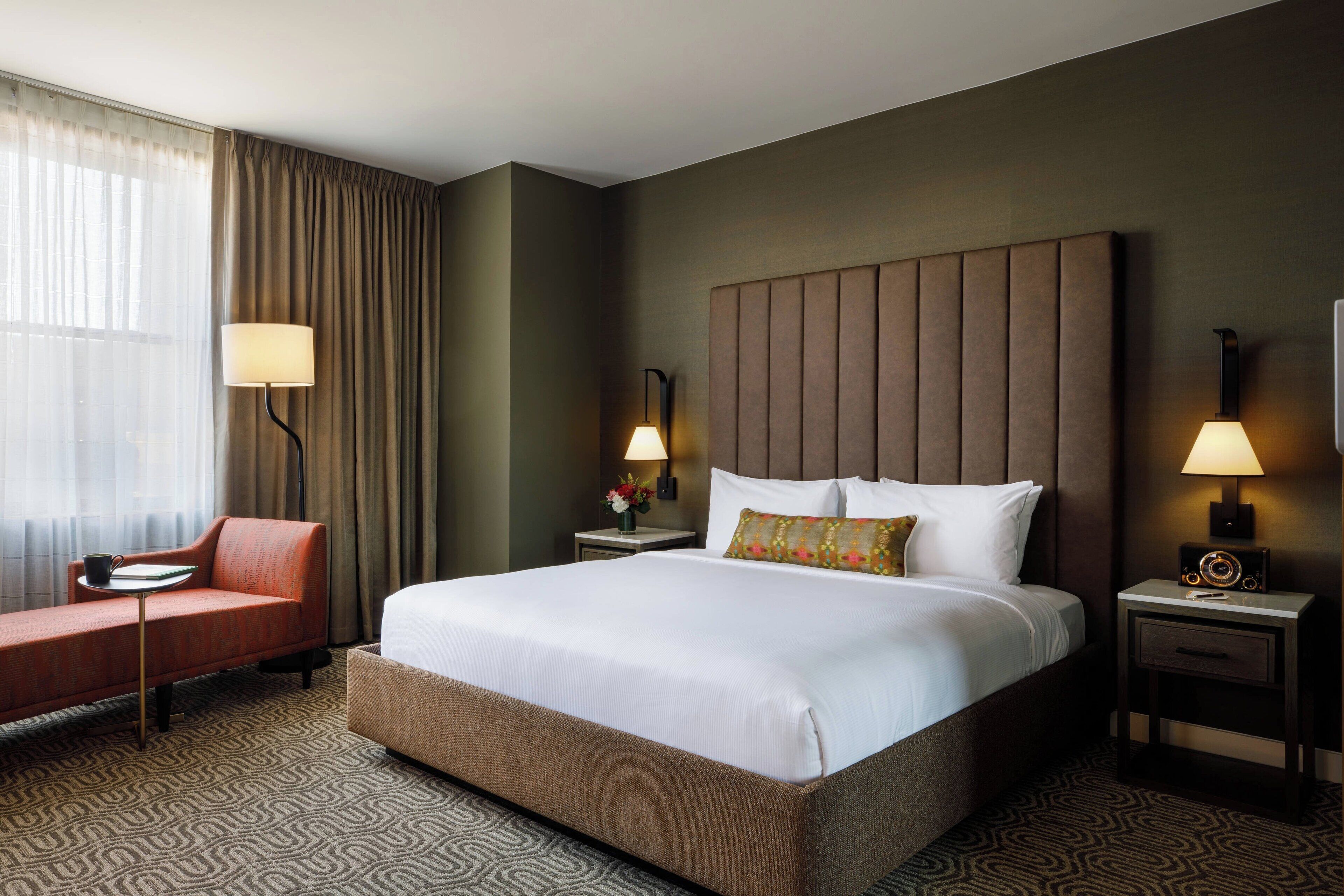 Habitación de hotel con una cama grande, un sofá, dos mesitas de noche con lámparas y una ventana con cortinas.