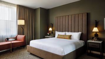 Habitación de hotel con una cama grande, un sofá, dos mesitas de noche con lámparas y una ventana con cortinas.