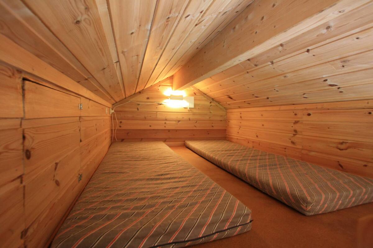 Ein kleiner, gemütlicher Raum mit Holzwänden und schrägverdeckter Decke. Es gibt zwei Betten mit gestreifter Bettwäsche.