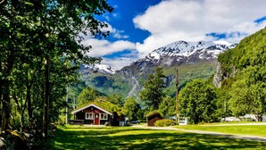 Uma cabana pequena com telhado vermelho e paredes brancas, rodeada por vegetação exuberante e montanhas ao fundo.