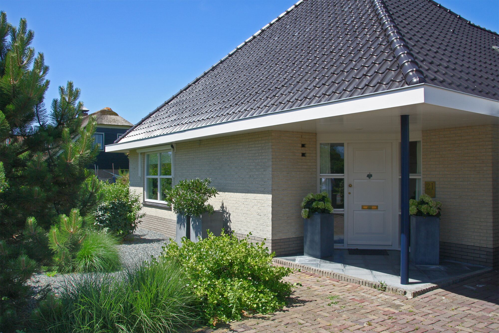 Une maison de plain-pied avec un toit recouvert de tuiles sombres, une porte d’entrée blanche et un petit porche agrémenté de plantes en pot.