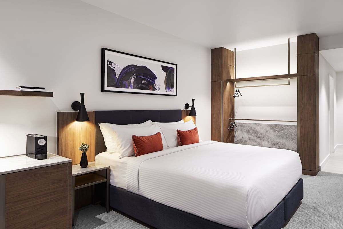 Een moderne hotelkamer met een groot bed, een nachtkastje met een lamp, een lijst met kunst aan de muur en ingebouwde houten kasten.