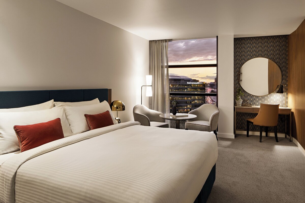Een hotelkamer met een groot bed, een bureau, een stoel, een ronde spiegel en uitzicht op een stadslandschap door een raam.