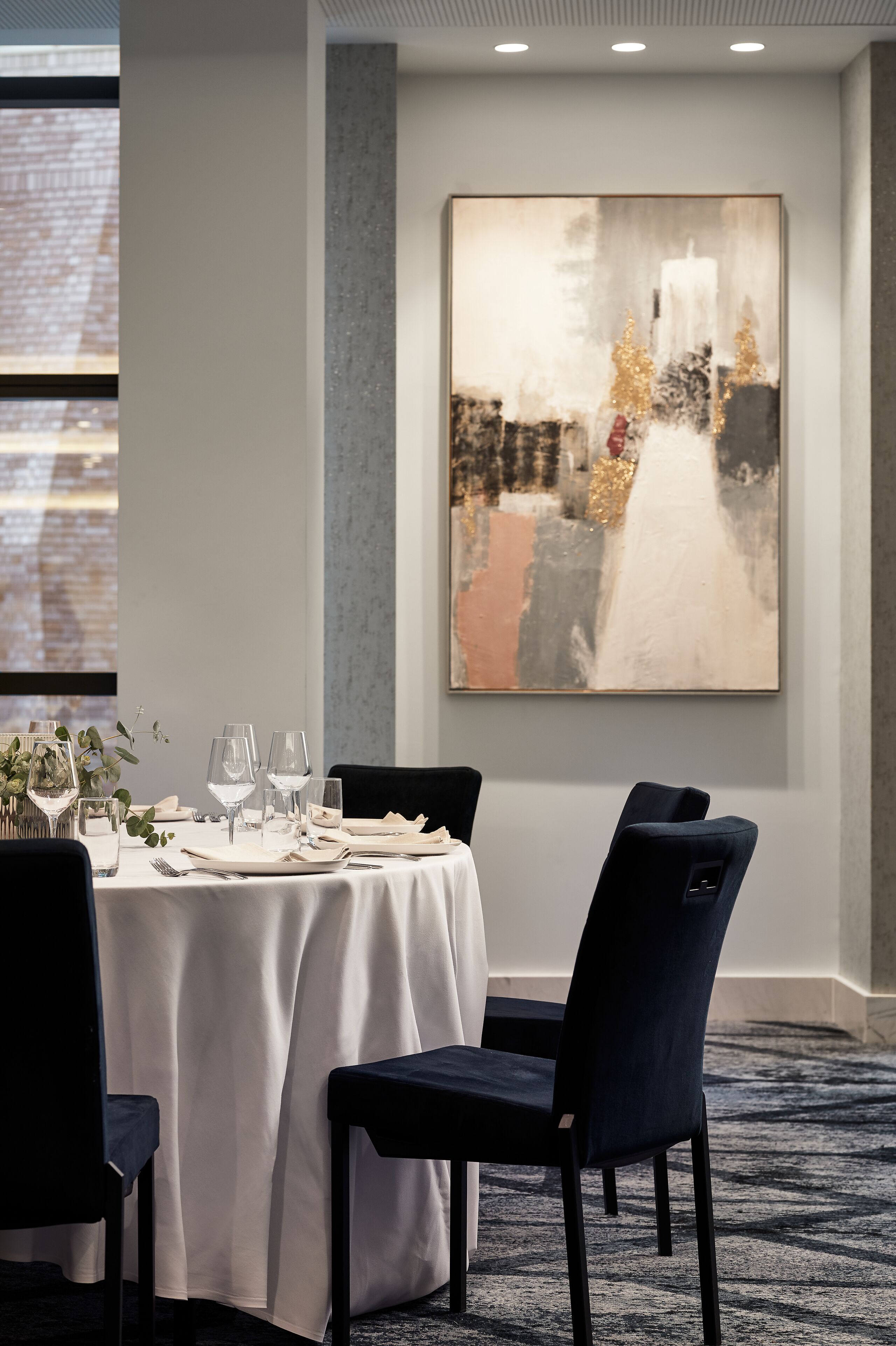 Un comedor con una mesa dispuesta para dos personas, un cuadro abstracto grande en la pared y vista a un edificio de ladrillo a través de una ventana.