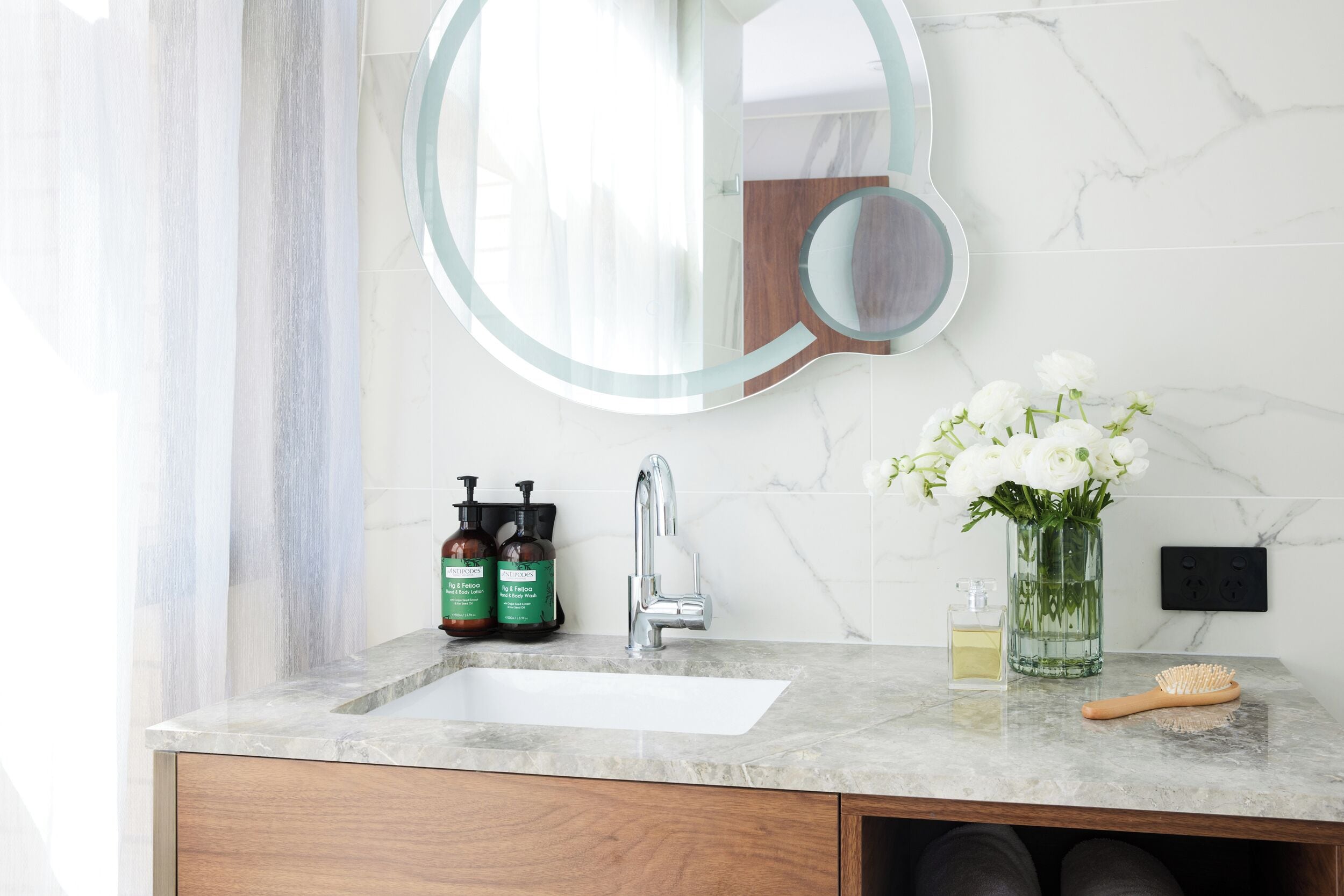 A bathroom with a marble sink, a round mirror, a vase with white flowers, and two bottles of shampoo.