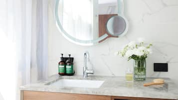 A bathroom with a marble sink, a round mirror, a vase with white flowers, and two bottles of shampoo.