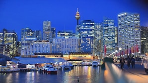 A waterfront cityscape with modern buildings, a prominent tower, and a marina with boats.