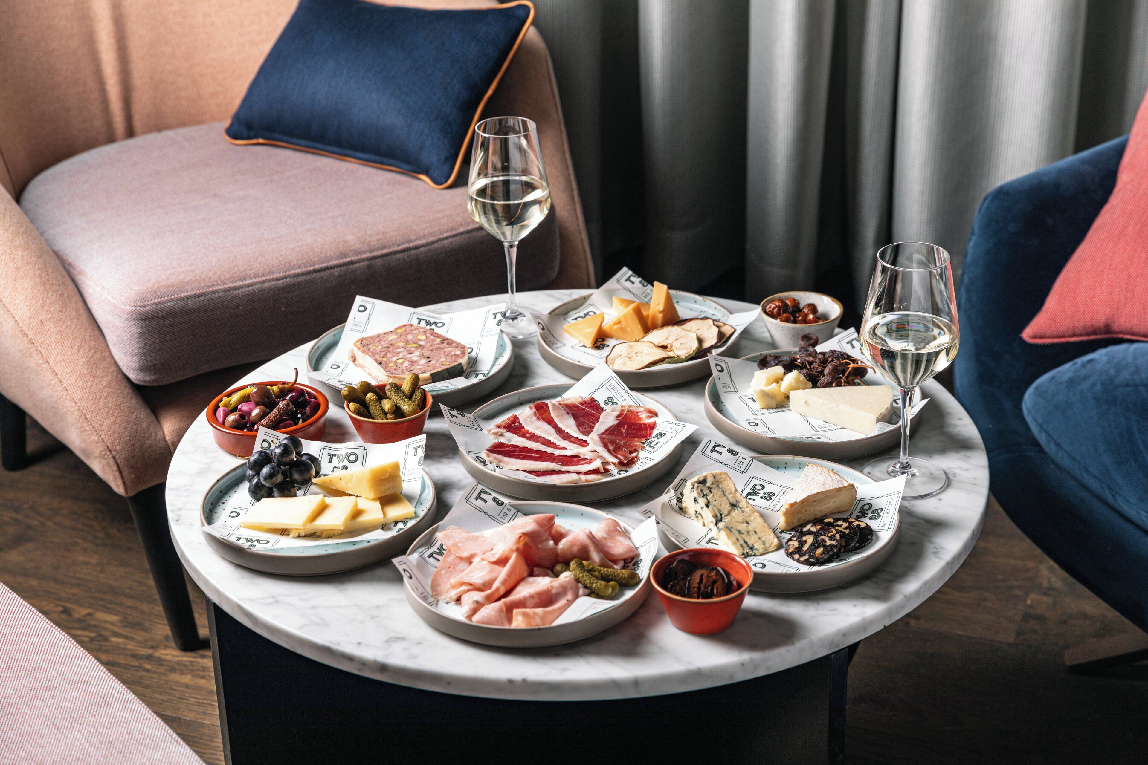 Una mesa de mármol con varios quesos, carnes y frutas, acompañada de copas de vino, en una habitación acogedora con una silla azul y rosa.