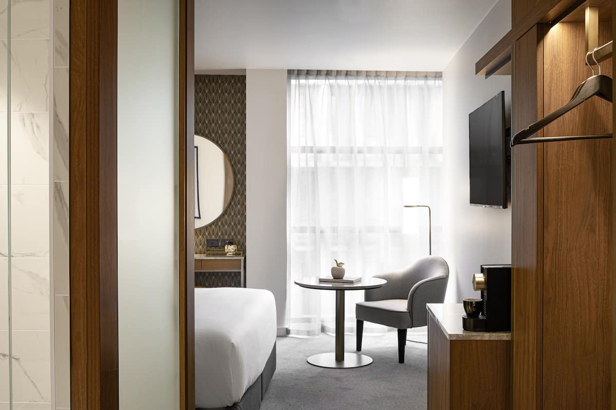 Een hotelkamer met een bed, een ronde tafel met een stoel, een koffieapparaat, een aan de muur bevestigde televisie en een raam met dunne gordijnen.