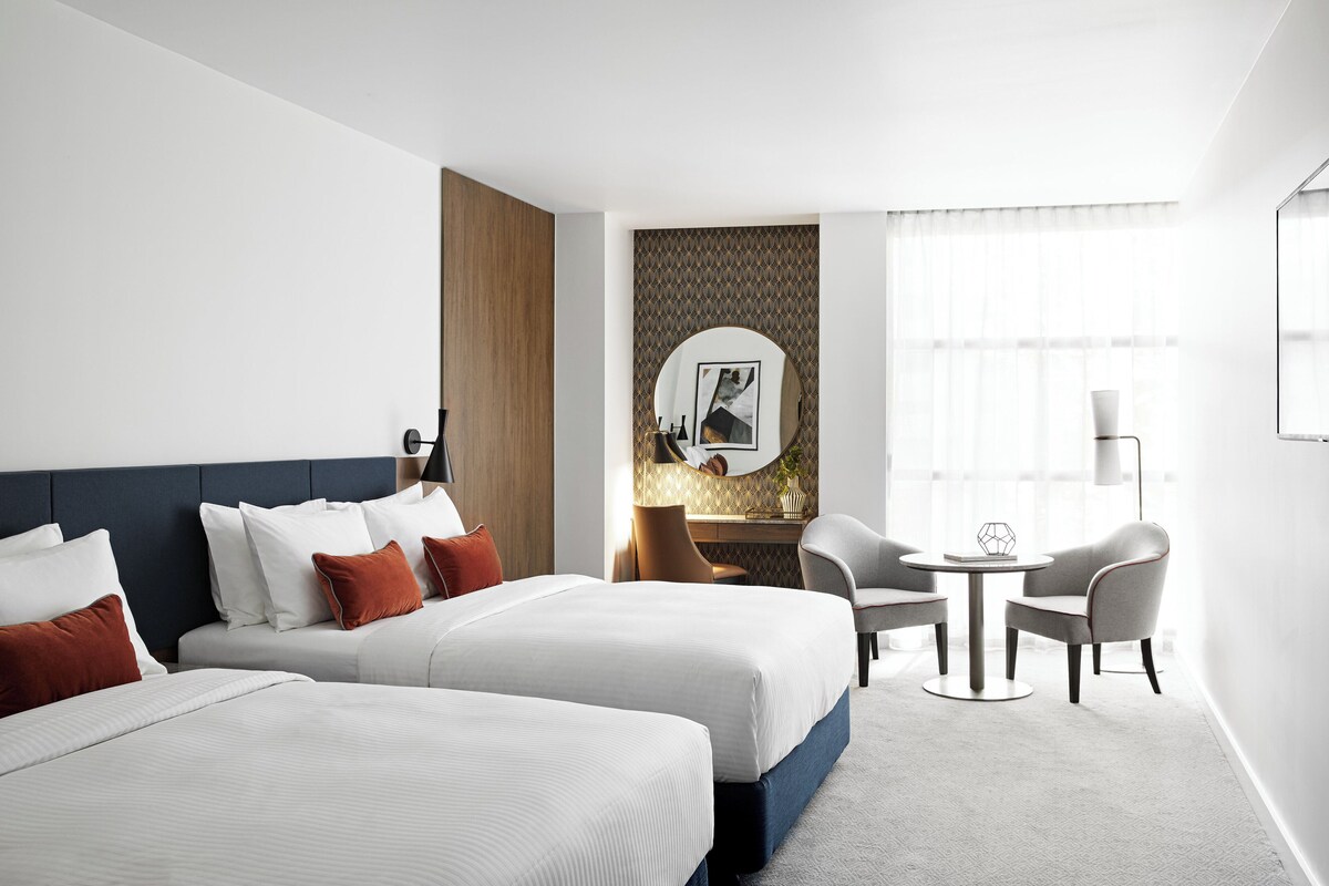 Een hotelkamer met twee bedden, een bureau, twee stoelen, een ronde tafel en een grote spiegel.