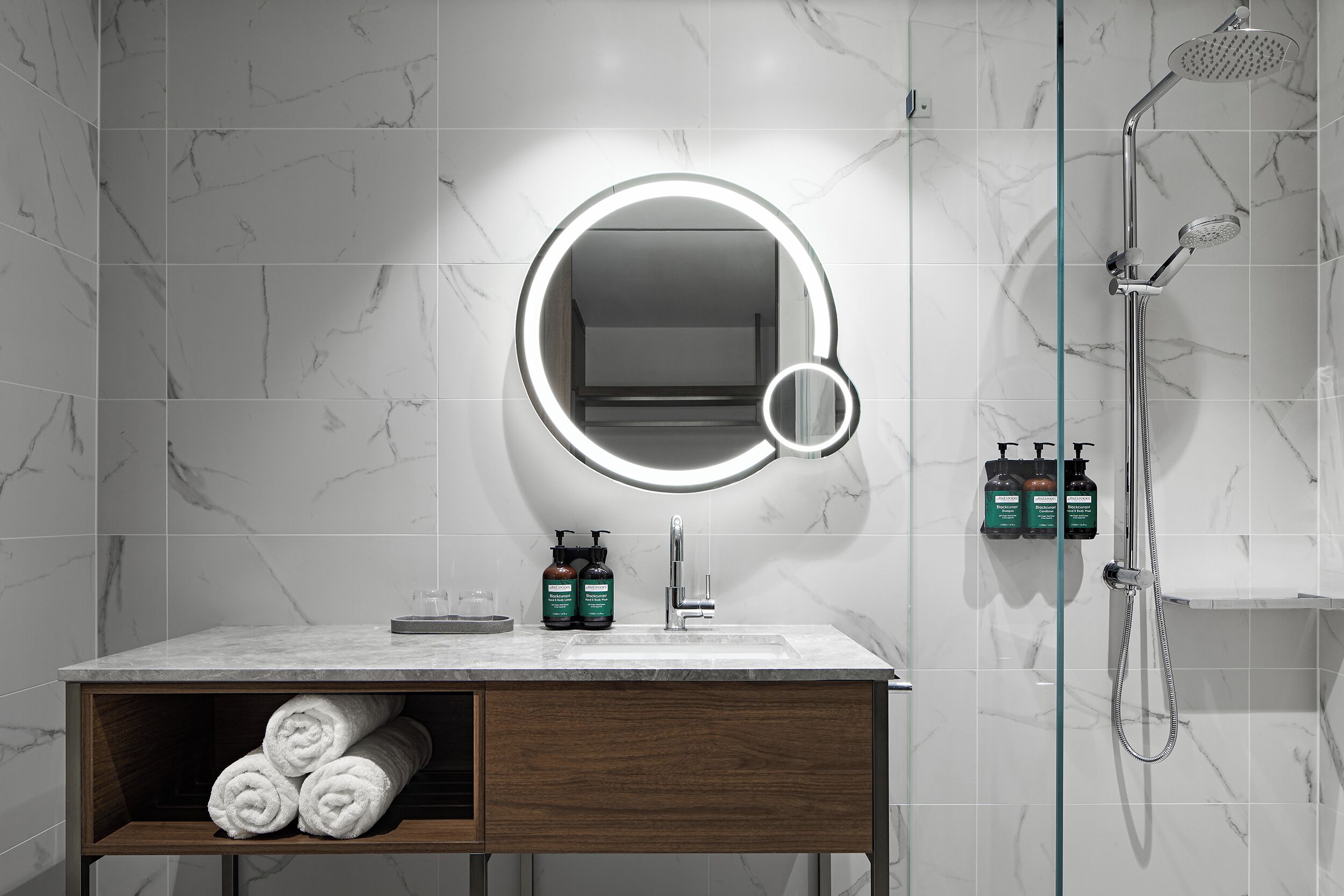 Un baño moderno con pared de azulejos de mármol, un espejo redondo con luz, ducha y tocador de madera con toallas.