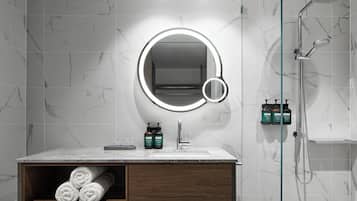 Un baño moderno con pared de azulejos de mármol, un espejo redondo con luz, ducha y tocador de madera con toallas.
