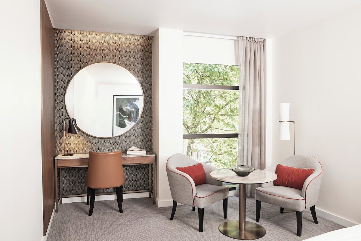 Hotelkamer met een ronde spiegel, een bureau, twee stoelen, een klein tafeltje en een raam met gordijnen.