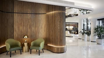 En modern entréhall med en böjd väggpanel i trä, två gröna fåtöljer, en guldfärgad lampa och krukväxter.