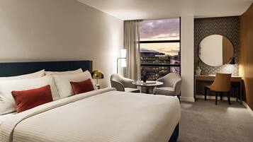 Quarto de hotel com uma cama grande, uma escrivaninha, duas cadeiras, uma mesa redonda, uma janela ampla com vista para a cidade e um espelho decorativo.
