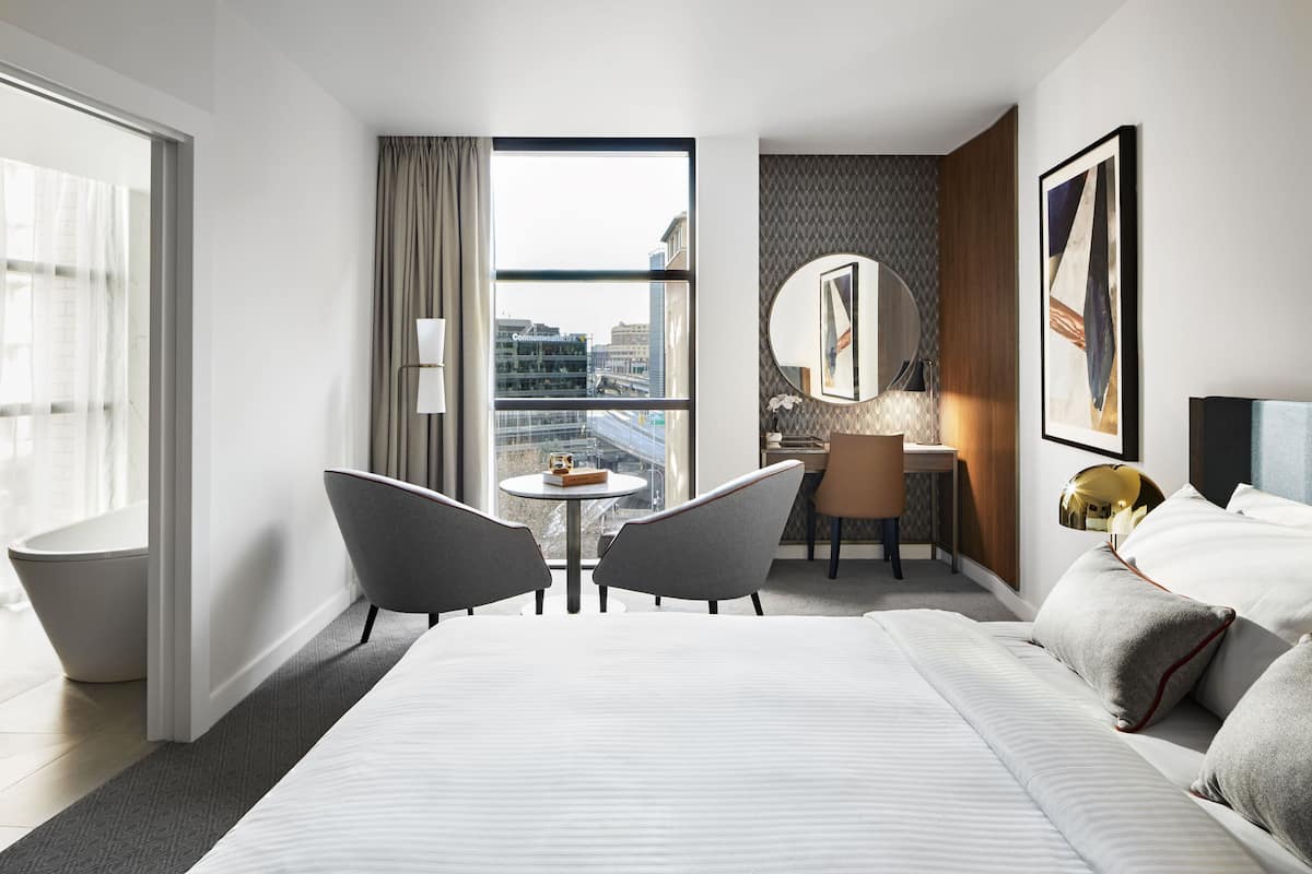 Een moderne hotelkamer met een groot bed, een bureau, twee stoelen, een ronde spiegel en uitzicht op de stad.