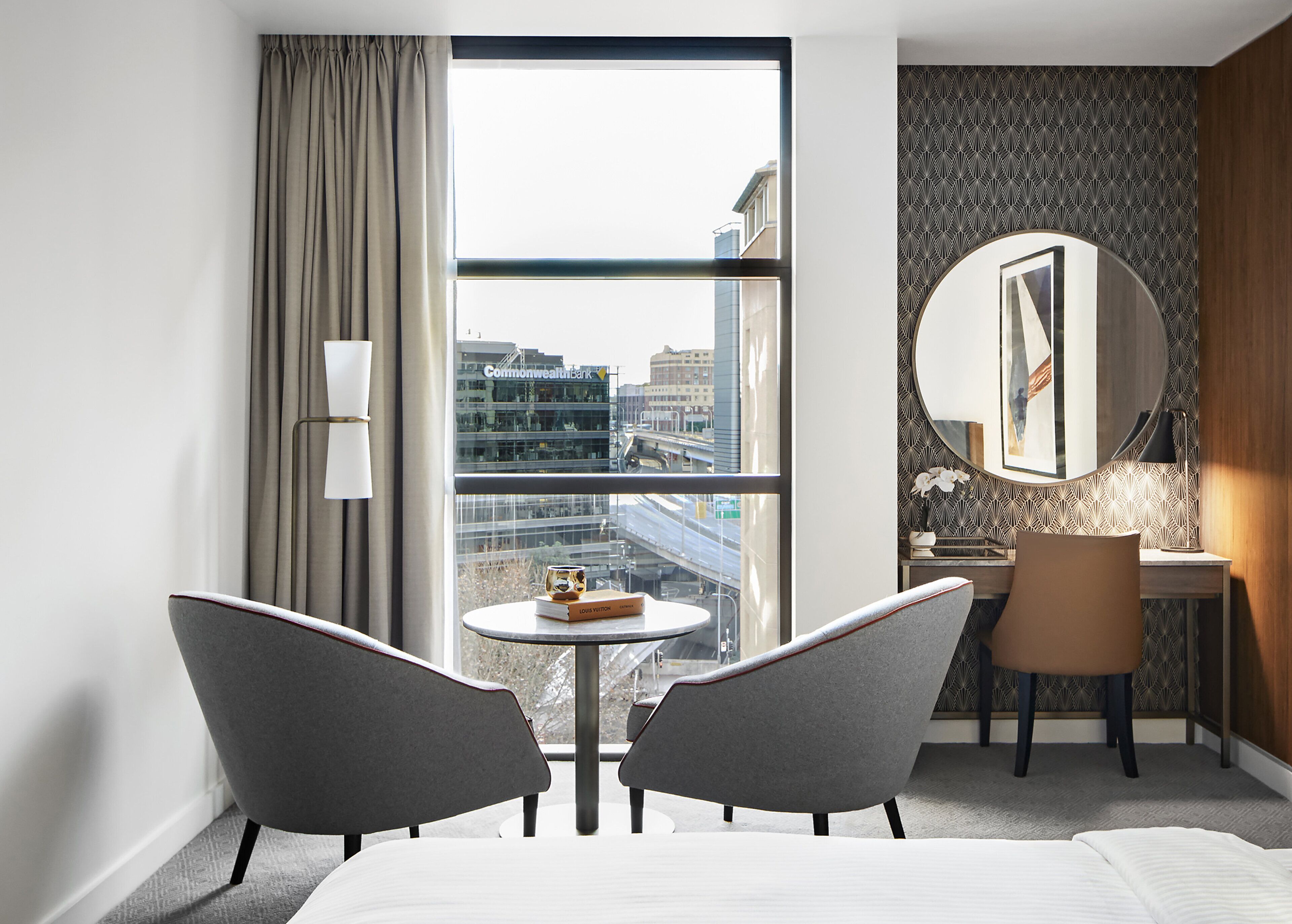 A hotel room with a large window, a round mirror, a desk, two chairs, and a bed.