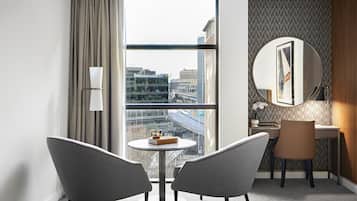 A hotel room with a large window, a round mirror, a desk, two chairs, and a bed.