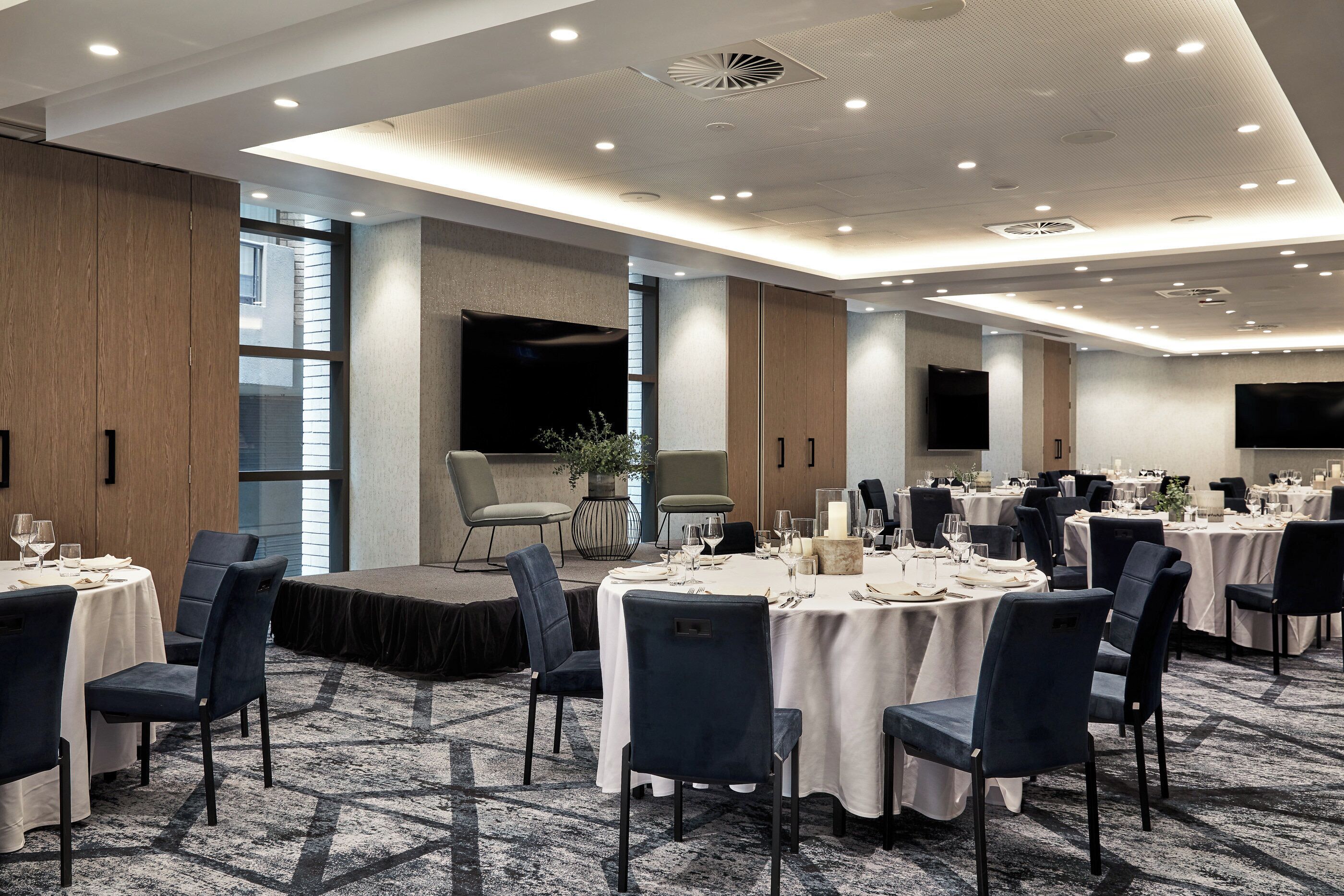 Un comedor de hotel con mesas redondas, sillas y un escenario para presentaciones.