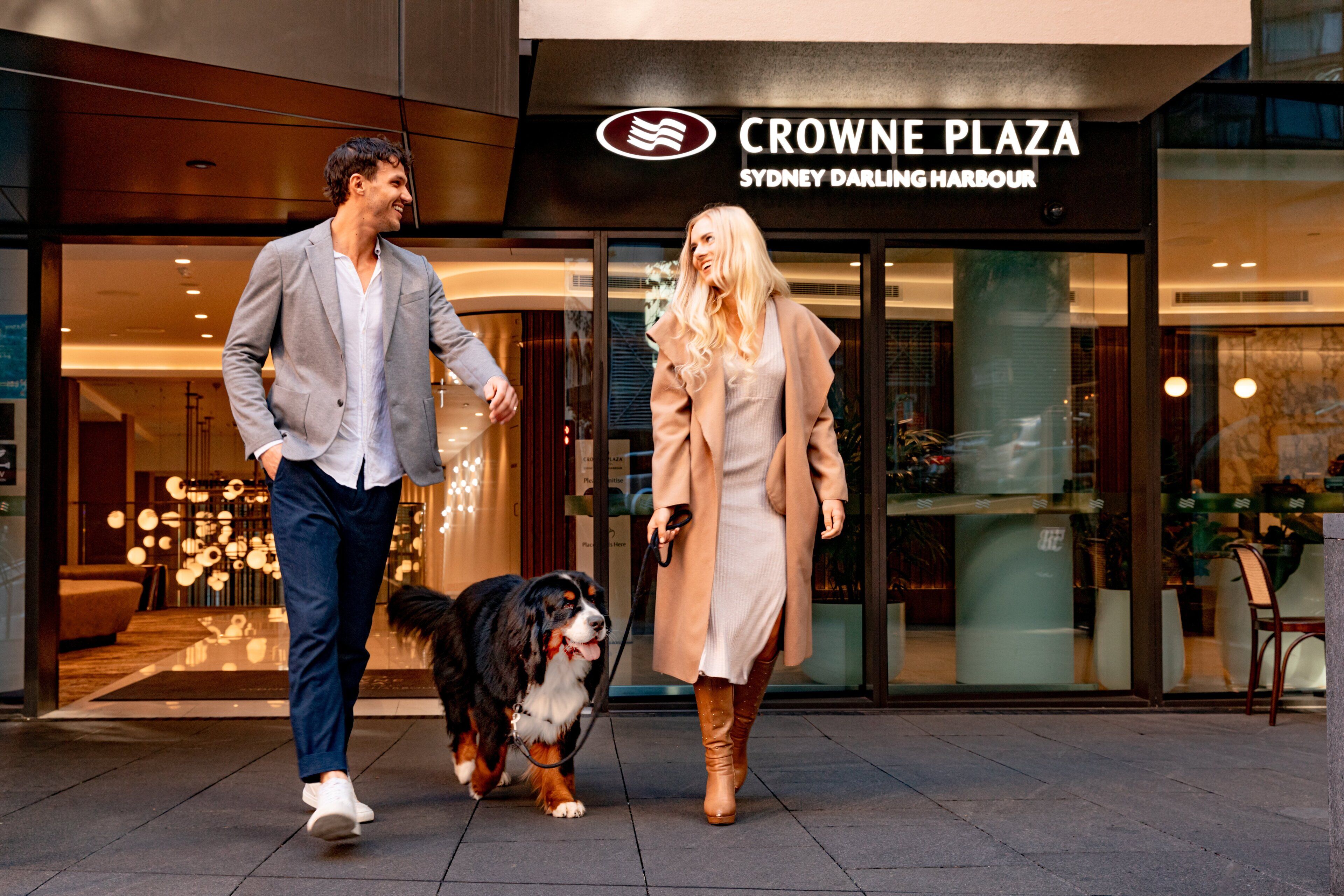 En man och en kvinna promenerar med en hund framför Crowne Plaza Sydney Darling Harbour.