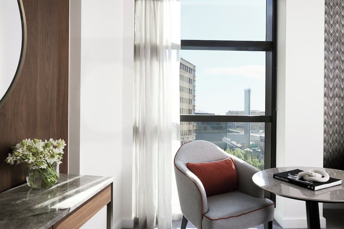 Een moderne kamer met uitzicht op een stadsbeeld, een grijze fauteuil met een rode kussens, een marmeren tafel met een boek en een wit object, en een vaas met witte bloemen op een marmeren oppervlak.