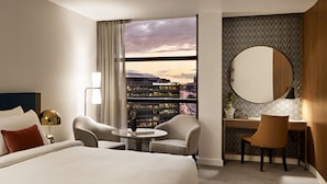 A hotel room with a large bed, two armchairs, a small round table, a desk with a chair, a large window with a view of a cityscape, and a round mirror on the wall.