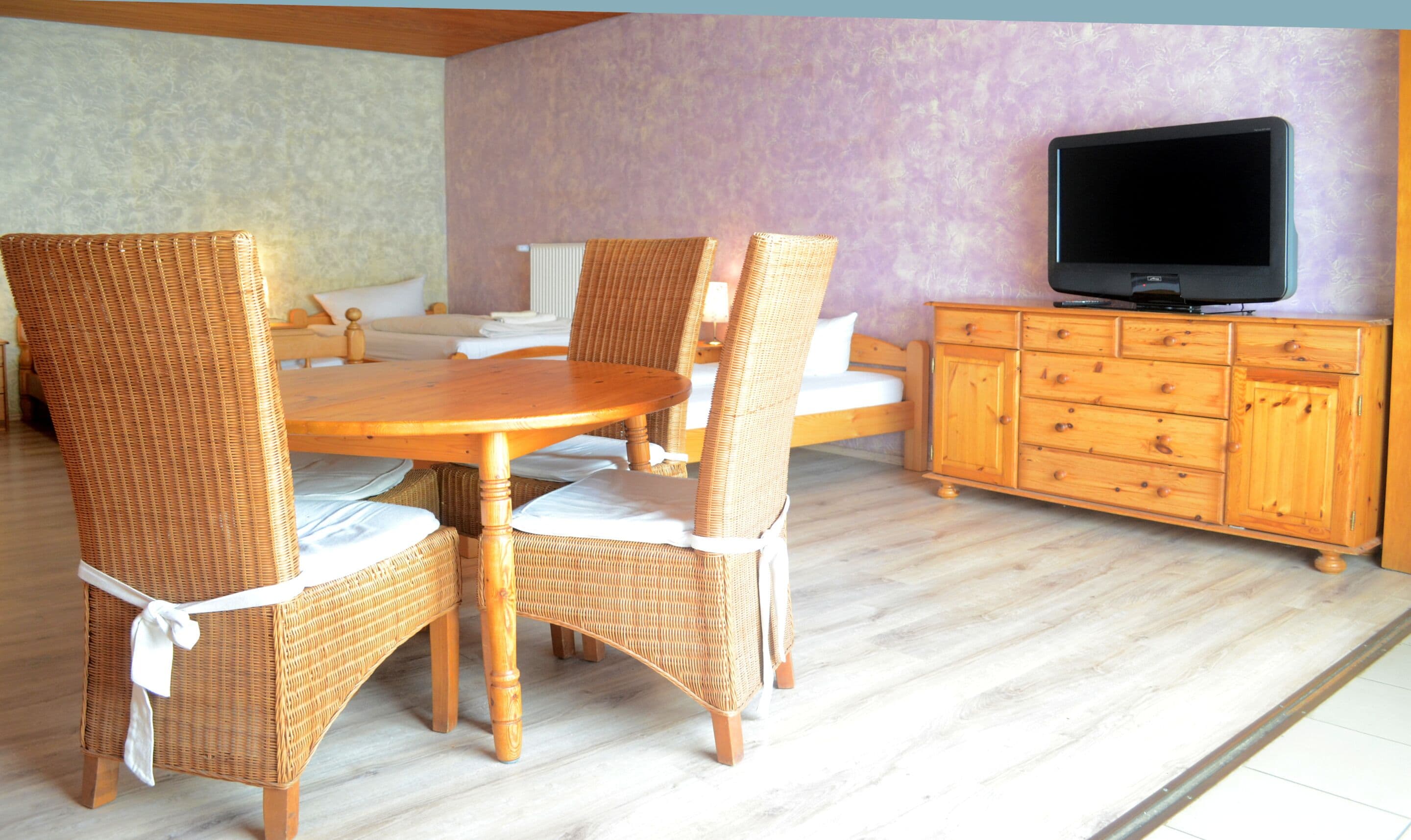Ein Essbereich mit Korbstühlen und einem Holztisch, einem Fernseher auf einer Holzkommode und einer violetten Wand im Hintergrund.
