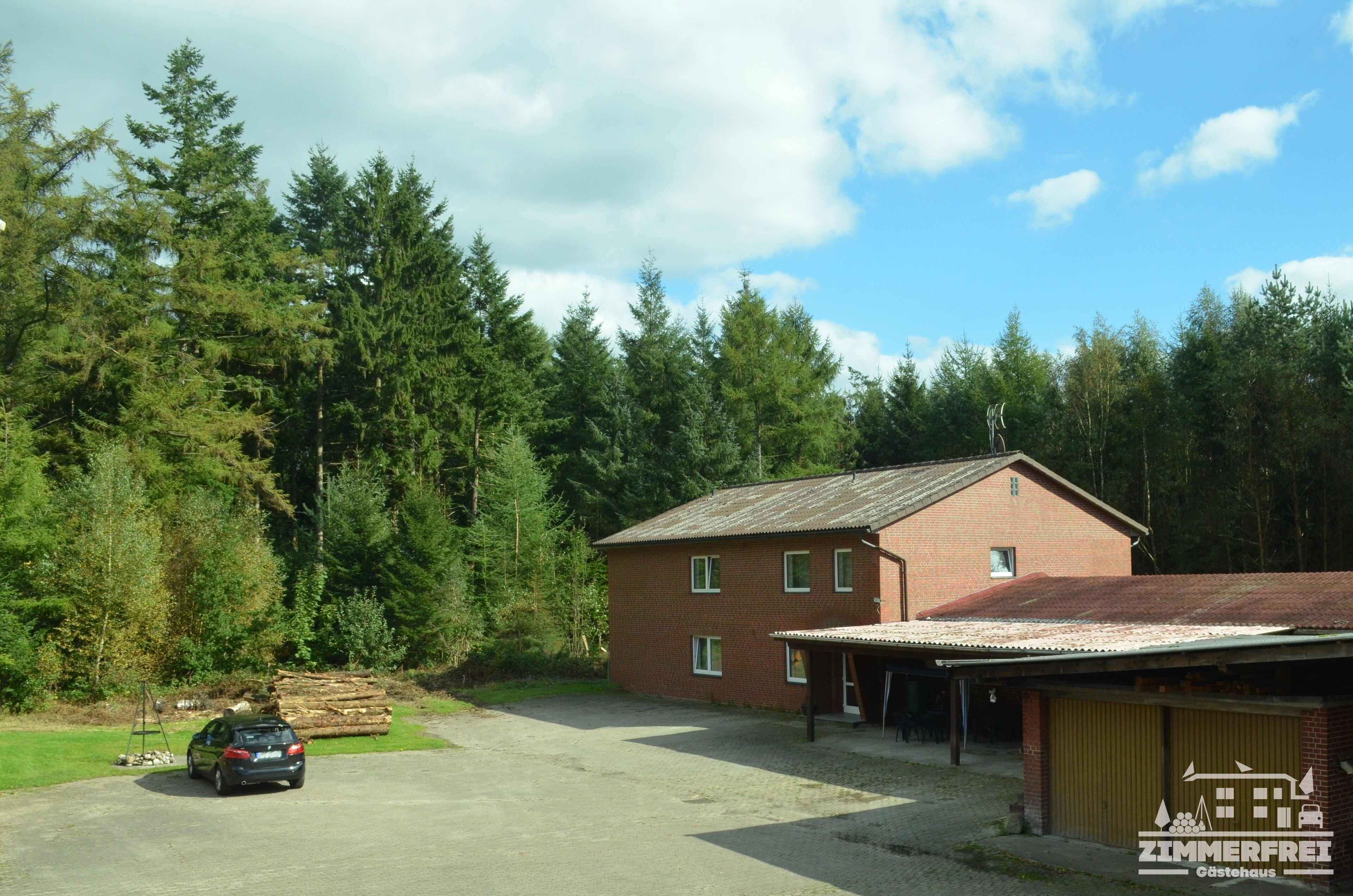 Paesaggio rurale con un edificio a due piani in mattoni, un'auto parcheggiata davanti e uno sfondo boschivo.