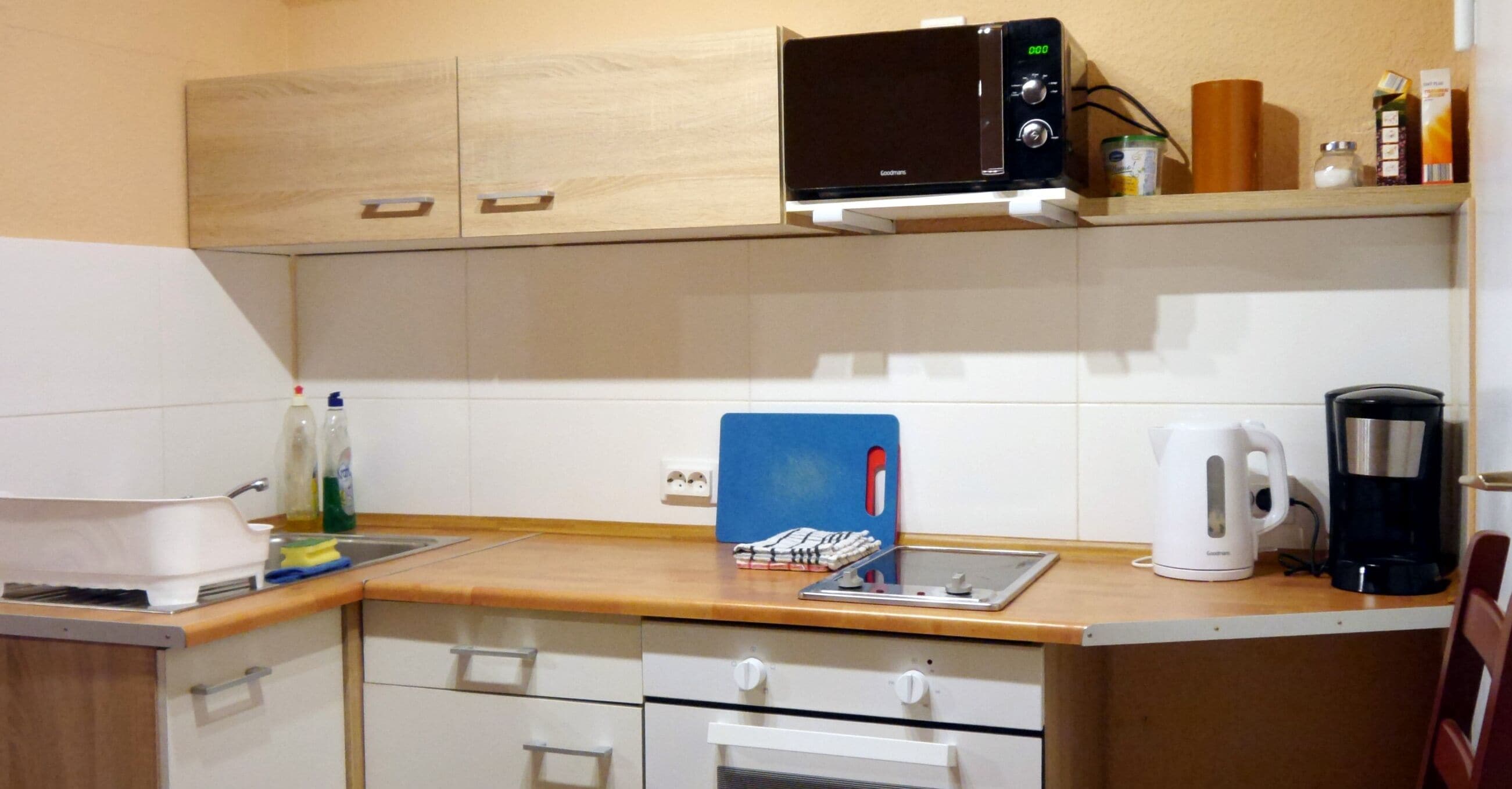 A kitchen with a microwave, kettle, coffee maker, and a cutting board on the counter.