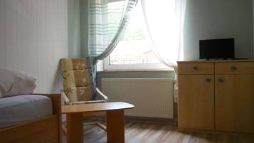 Une pièce comprenant un lit, une chaise en bois, une petite table, un téléviseur posé sur un meuble en bois et une fenêtre avec des rideaux.